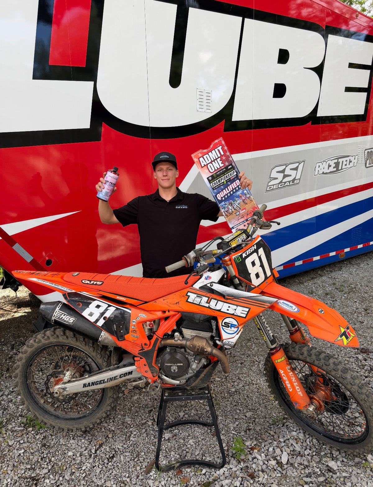 Motocross rider in an Enraged shirt holding a bottle beside an orange dirt bike at a race trailer.